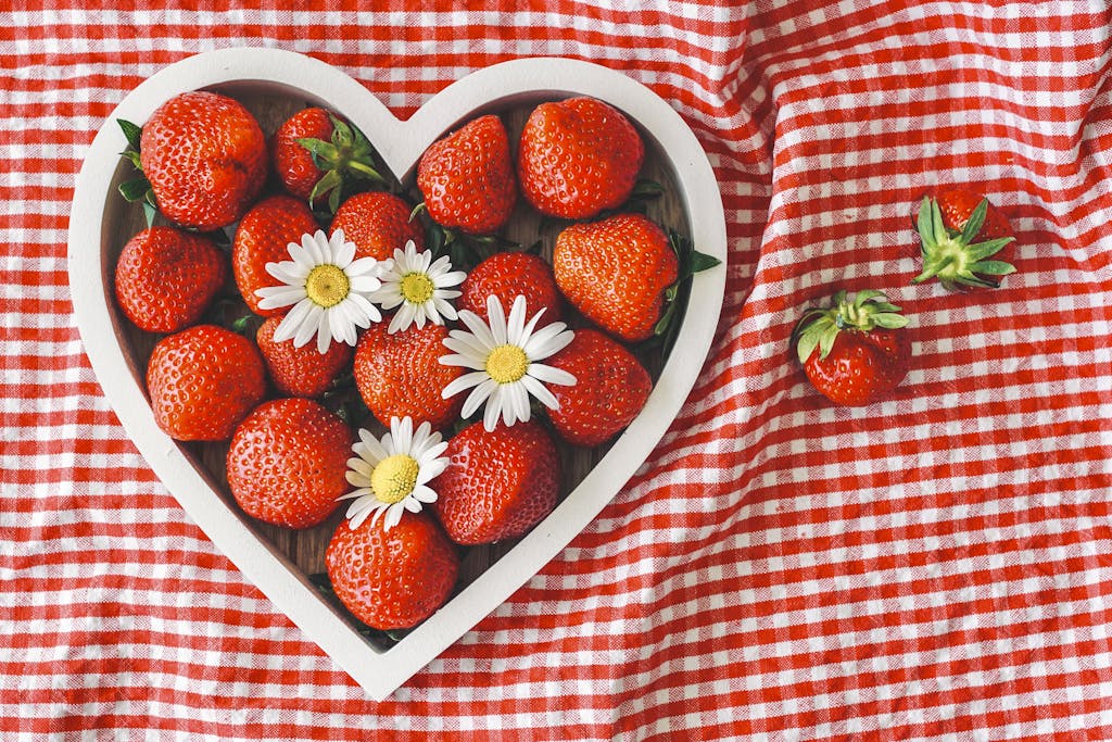Fresh strawberries and daisies in a heart-shaped bowl on a red checkered cloth.